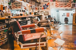 Interior of a barber shop featuring multiple vintage barber chairs lined up along a wooden floor. The walls are adorned with various signs, photos, and memorabilia, and shelves are stocked with grooming products. The space has a nostalgic and classic atmosphere.