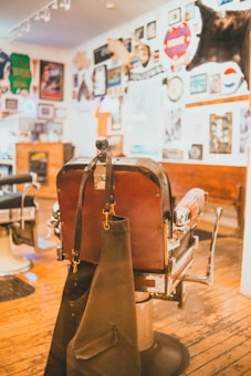 A vintage barber chair with leather upholstery and metal accents sits on a wooden floor. An apron is draped over the back of the chair. The walls are adorned with various memorabilia, such as framed photos, signs, and taxidermy, contributing to a nostalgic and eclectic atmosphere.