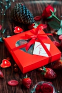 A bright red gift box adorned with a ribbon and a card is surrounded by strawberries, a pine cone, red roses, and heart-shaped candles on a wooden surface.