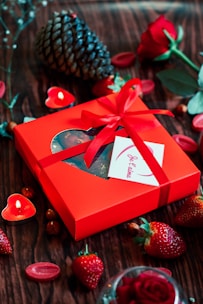 A bright red gift box adorned with a ribbon and a card is surrounded by strawberries, a pine cone, red roses, and heart-shaped candles on a wooden surface.