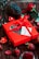 A bright red gift box adorned with a ribbon and a card is surrounded by strawberries, a pine cone, red roses, and heart-shaped candles on a wooden surface.