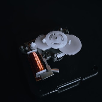 A close-up view of a mechanical gear system with several interlocking white gears and a copper coil. The components are mounted on a dark background that emphasizes the intricate details of the machinery.