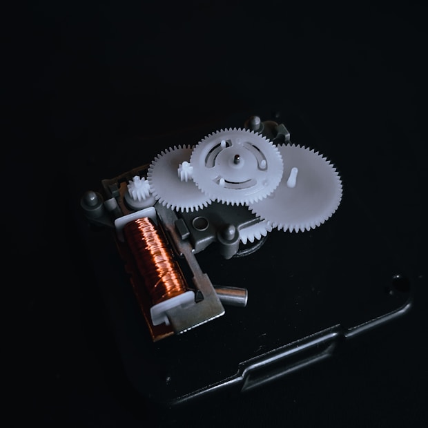 A close-up view of a mechanical gear system with several interlocking white gears and a copper coil. The components are mounted on a dark background that emphasizes the intricate details of the machinery.