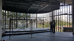 A construction site with an incomplete structure, featuring scaffolding and two people in hard hats standing together, possibly discussing plans. The background shows greenery and partially built surroundings.
