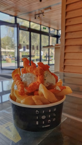 A bowl of crispy fries topped with a creamy sauce and crunchy orange snacks is placed on a glass table. The setting is a modern, well-lit cafe with large glass windows and wooden paneling, offering a view of the outdoors.