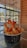A bowl of crispy fries topped with a creamy sauce and crunchy orange snacks is placed on a glass table. The setting is a modern, well-lit cafe with large glass windows and wooden paneling, offering a view of the outdoors.