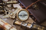 A collection of vintage nautical instruments arranged on a textured surface, including a brass compass, sextant, and an old telescope. These items rest on a burlap cloth alongside a leather-bound journal with a decorative ship wheel charm.