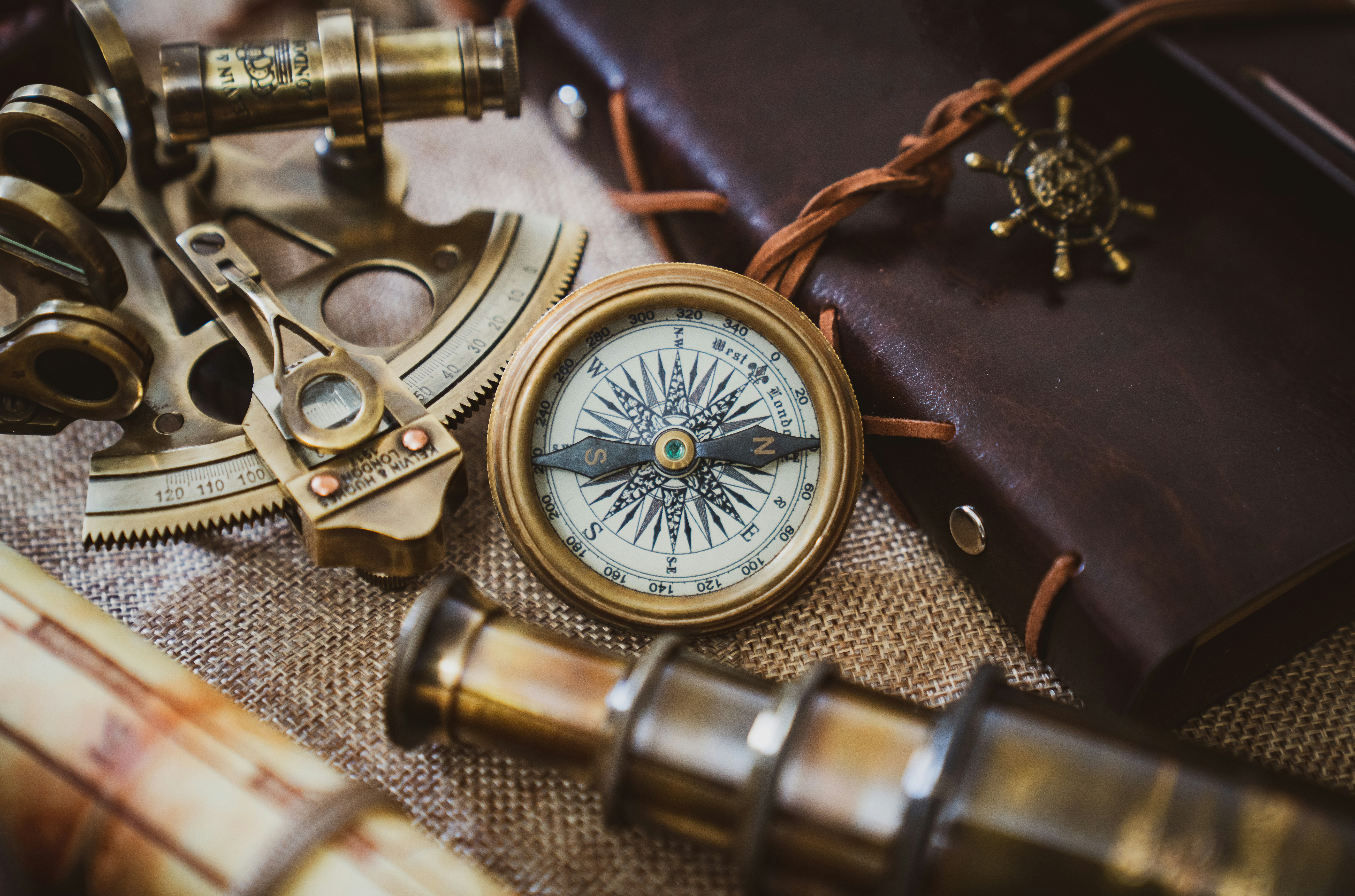 A collection of vintage nautical instruments arranged on a textured surface, including a brass compass, sextant, and an old telescope. These items rest on a burlap cloth alongside a leather-bound journal with a decorative ship wheel charm.