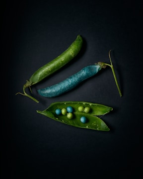 Three pea pods are arranged on a dark background, with one pod open to reveal green and blue peas inside. The pods vary in color, with one appearing naturally green and another in an unusual blue shade.