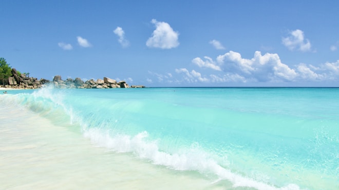 A pristine beach with turquoise waves gently crashing on the sandy shore under a clear blue sky. Fluffy white clouds are scattered across the horizon and rocky formations are visible in the distance.