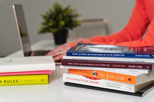 Several books are stacked on a desk, along with a pair of hands typing on a laptop in the background. The books have various colorful covers, and a small plant is visible in the background.