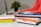 Several books are stacked on a desk, along with a pair of hands typing on a laptop in the background. The books have various colorful covers, and a small plant is visible in the background.