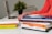 Several books are stacked on a desk, along with a pair of hands typing on a laptop in the background. The books have various colorful covers, and a small plant is visible in the background.