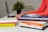Several books are stacked on a desk, along with a pair of hands typing on a laptop in the background. The books have various colorful covers, and a small plant is visible in the background.