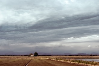 A vast agricultural landscape with fields stretching towards the horizon under heavy, overcast skies. In the midground, a solitary small building stands next to a tree. Mountains are faintly visible in the distance, beneath the thick, layered clouds that suggest an impending storm.