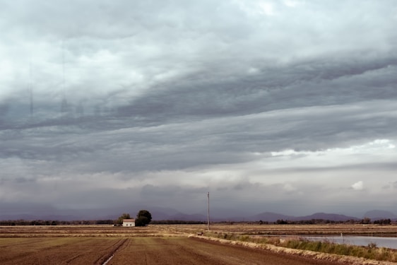 A vast agricultural landscape with fields stretching towards the horizon under heavy, overcast skies. In the midground, a solitary small building stands next to a tree. Mountains are faintly visible in the distance, beneath the thick, layered clouds that suggest an impending storm.