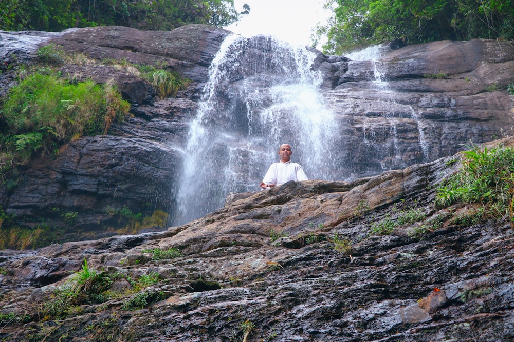 man in white shirt sitting on brown rock near waterfalls during daytime