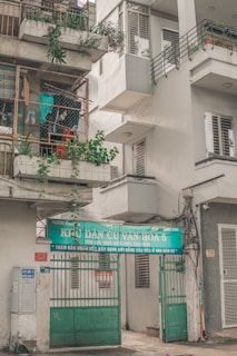 An urban residential building with a sign over the entrance featuring Vietnamese text. The building has balconies with potted plants and a collection of hanging clothes within a wire enclosure. Various elements like air conditioning units and electrical wires are visible, contributing to a lived-in appearance.