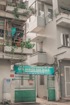 An urban residential building with a sign over the entrance featuring Vietnamese text. The building has balconies with potted plants and a collection of hanging clothes within a wire enclosure. Various elements like air conditioning units and electrical wires are visible, contributing to a lived-in appearance.