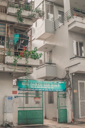 An urban residential building with a sign over the entrance featuring Vietnamese text. The building has balconies with potted plants and a collection of hanging clothes within a wire enclosure. Various elements like air conditioning units and electrical wires are visible, contributing to a lived-in appearance.