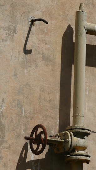 A weathered metal valve attached to an old pipe, mounted on a rough, beige-colored concrete wall. The shadows of the valve and pipe create stark contrasts on the wall.