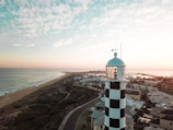 A black and white striped lighthouse overlooks the coastline at sunset. The scene includes a beach extending into the horizon, ocean waves gently rolling in, and a cluster of residential houses near the shore. The sky is partly cloudy with hues of pink and blue, adding a serene backdrop.