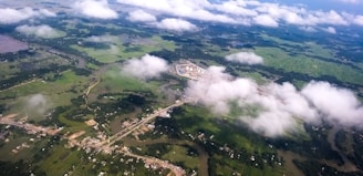 An aerial view of a vast landscape featuring lush green fields, meandering rivers, and scattered clouds casting shadows on the ground below. A small cluster of buildings is visible in the center, with roads and infrastructure connecting around it.
