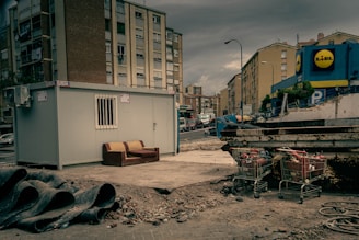 An urban setting with a construction site featuring a portable cabin with a barred window. A worn-out couch is placed next to the cabin on a patch of concrete. Nearby, construction pipes are stacked, and there are several shopping carts filled with debris. Apartment buildings border the background, with a commercial building visible on the right displaying a store logo.