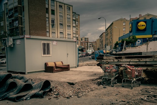 An urban setting with a construction site featuring a portable cabin with a barred window. A worn-out couch is placed next to the cabin on a patch of concrete. Nearby, construction pipes are stacked, and there are several shopping carts filled with debris. Apartment buildings border the background, with a commercial building visible on the right displaying a store logo.