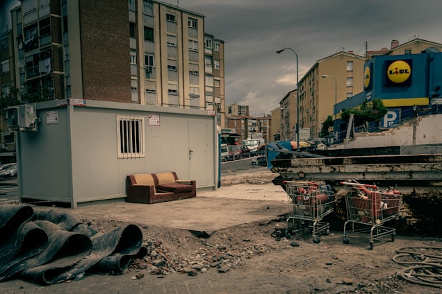 An urban setting with a construction site featuring a portable cabin with a barred window. A worn-out couch is placed next to the cabin on a patch of concrete. Nearby, construction pipes are stacked, and there are several shopping carts filled with debris. Apartment buildings border the background, with a commercial building visible on the right displaying a store logo.