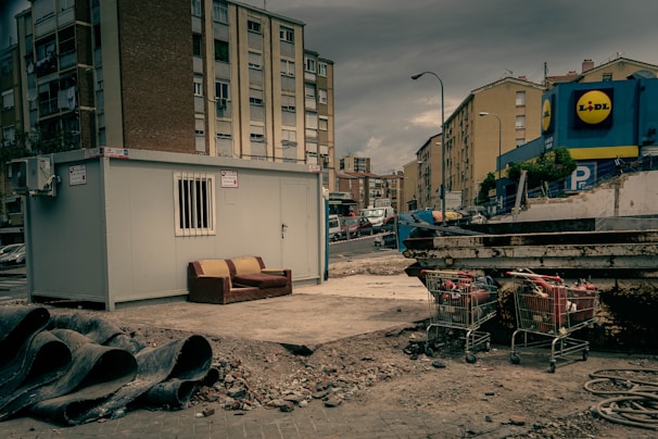 An urban setting with a construction site featuring a portable cabin with a barred window. A worn-out couch is placed next to the cabin on a patch of concrete. Nearby, construction pipes are stacked, and there are several shopping carts filled with debris. Apartment buildings border the background, with a commercial building visible on the right displaying a store logo.