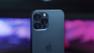 A modern smartphone with a sleek design, featuring multiple camera lenses on the back. The phone has a smooth, dark finish, and there is a blurred, vibrant background in shades of purple and pink.