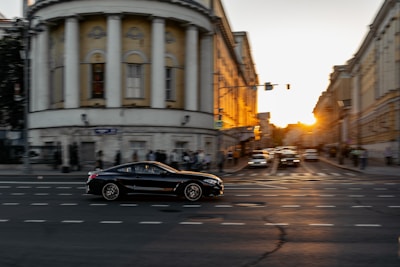 A sleek car driving through an urban street during sunset with city buildings visible.