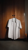 A neatly ironed white shirt is hanging on a wooden wardrobe door. The wardrobe has a textured, natural wood finish with subtle lighting creating a warm atmosphere. A hanger supports the shirt, and a small detergent bottle can be seen partially in the corner.