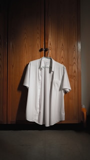 A neatly ironed white shirt is hanging on a wooden wardrobe door. The wardrobe has a textured, natural wood finish with subtle lighting creating a warm atmosphere. A hanger supports the shirt, and a small detergent bottle can be seen partially in the corner.