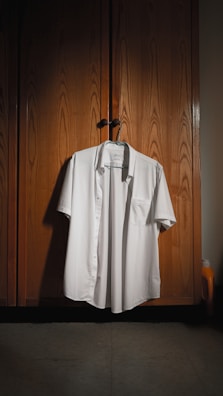 A neatly ironed white shirt is hanging on a wooden wardrobe door. The wardrobe has a textured, natural wood finish with subtle lighting creating a warm atmosphere. A hanger supports the shirt, and a small detergent bottle can be seen partially in the corner.