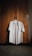 A neatly ironed white shirt is hanging on a wooden wardrobe door. The wardrobe has a textured, natural wood finish with subtle lighting creating a warm atmosphere. A hanger supports the shirt, and a small detergent bottle can be seen partially in the corner.
