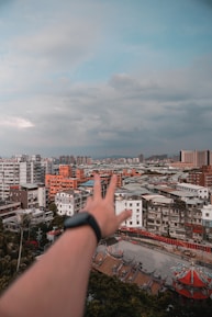 A cityscape with dense urban buildings, including high-rise apartments and traditional architectural details at the bottom. A blurred hand with a smartwatch in the foreground reaches towards the skyline under a partly cloudy sky.