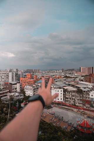A cityscape with dense urban buildings, including high-rise apartments and traditional architectural details at the bottom. A blurred hand with a smartwatch in the foreground reaches towards the skyline under a partly cloudy sky.