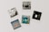 A collection of five polaroid photographs arranged on a flat surface. The top-left photo features a monochrome image with shelves and various objects. The top-middle shows a street view of a building with a sign that reads 'PHOTO'. The top-right displays a dark image with a bright rectangle in the center, possibly a light or reflection. The bottom-left shows a close-up of shutters with a sign that has an orange section. The bottom-center photo is a vibrant, abstract graffiti artwork in shades of blue and yellow.