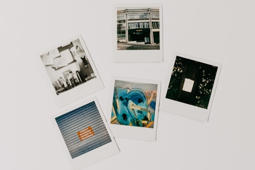 A collection of five polaroid photographs arranged on a flat surface. The top-left photo features a monochrome image with shelves and various objects. The top-middle shows a street view of a building with a sign that reads 'PHOTO'. The top-right displays a dark image with a bright rectangle in the center, possibly a light or reflection. The bottom-left shows a close-up of shutters with a sign that has an orange section. The bottom-center photo is a vibrant, abstract graffiti artwork in shades of blue and yellow.