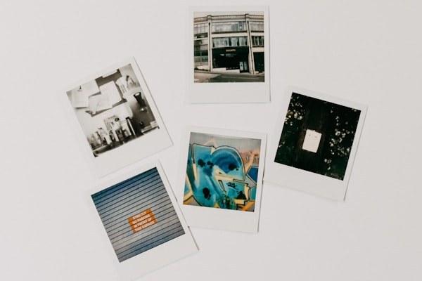 A collection of five polaroid photographs arranged on a flat surface. The top-left photo features a monochrome image with shelves and various objects. The top-middle shows a street view of a building with a sign that reads 'PHOTO'. The top-right displays a dark image with a bright rectangle in the center, possibly a light or reflection. The bottom-left shows a close-up of shutters with a sign that has an orange section. The bottom-center photo is a vibrant, abstract graffiti artwork in shades of blue and yellow.