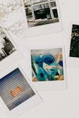 A collection of Polaroid photographs laid out on a white background. One features a building with the word 'PRONTO' on it, another shows colorful graffiti with blue and yellow hues, and a third displays a garage door with a sign that reads 'STRICTLY NO PARKING IN FRONT OF THIS GARAGE.' Dried flowers are visible in the top left corner, adding an artistic touch.
