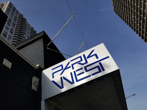 A modern architectural scene featuring an angular building with a large blue sign that reads 'Park West' in a stylized font. The building is constructed with dark materials, and the surrounding area includes other high-rise buildings against a clear blue sky.