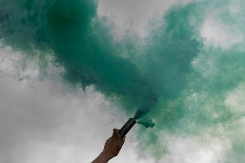 A hand holding a canister from which green smoke is billowing out against a grayish cloudy sky.