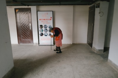 An elderly person is wearing a red coat and using a walking stick, standing in an indoor hallway area. The setting includes a framed poster on the wall with text and images as part of a display, along with two doors on either side of the person.