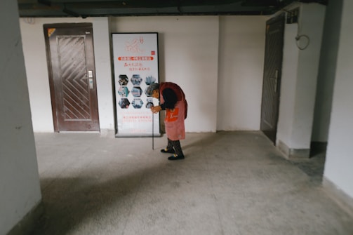An elderly person is wearing a red coat and using a walking stick, standing in an indoor hallway area. The setting includes a framed poster on the wall with text and images as part of a display, along with two doors on either side of the person.