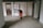 An elderly person is wearing a red coat and using a walking stick, standing in an indoor hallway area. The setting includes a framed poster on the wall with text and images as part of a display, along with two doors on either side of the person.