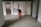 An elderly person is wearing a red coat and using a walking stick, standing in an indoor hallway area. The setting includes a framed poster on the wall with text and images as part of a display, along with two doors on either side of the person.
