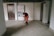 An elderly person is wearing a red coat and using a walking stick, standing in an indoor hallway area. The setting includes a framed poster on the wall with text and images as part of a display, along with two doors on either side of the person.